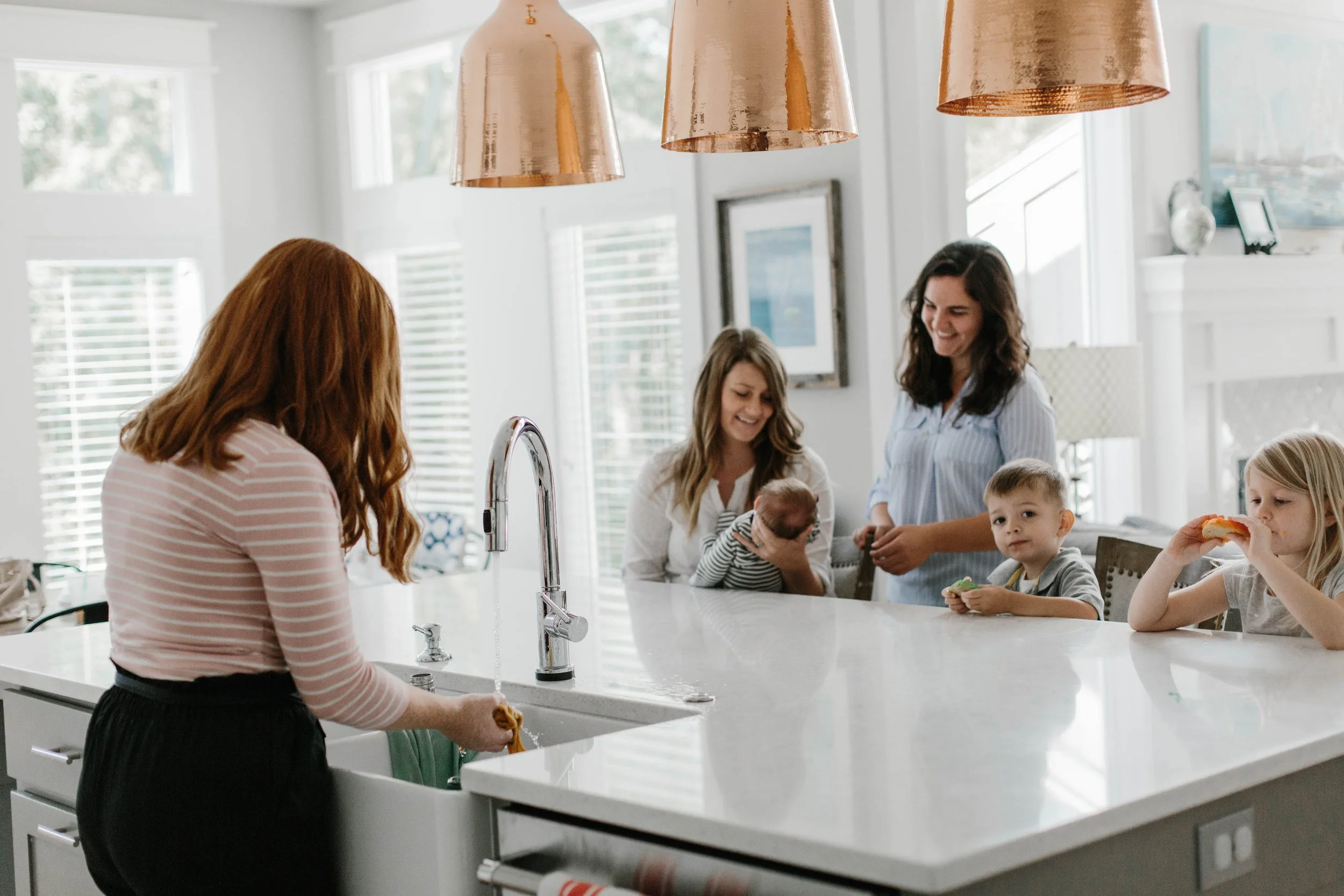 Parent holding newborn near kitchen island while children sit and another adult washes dishes