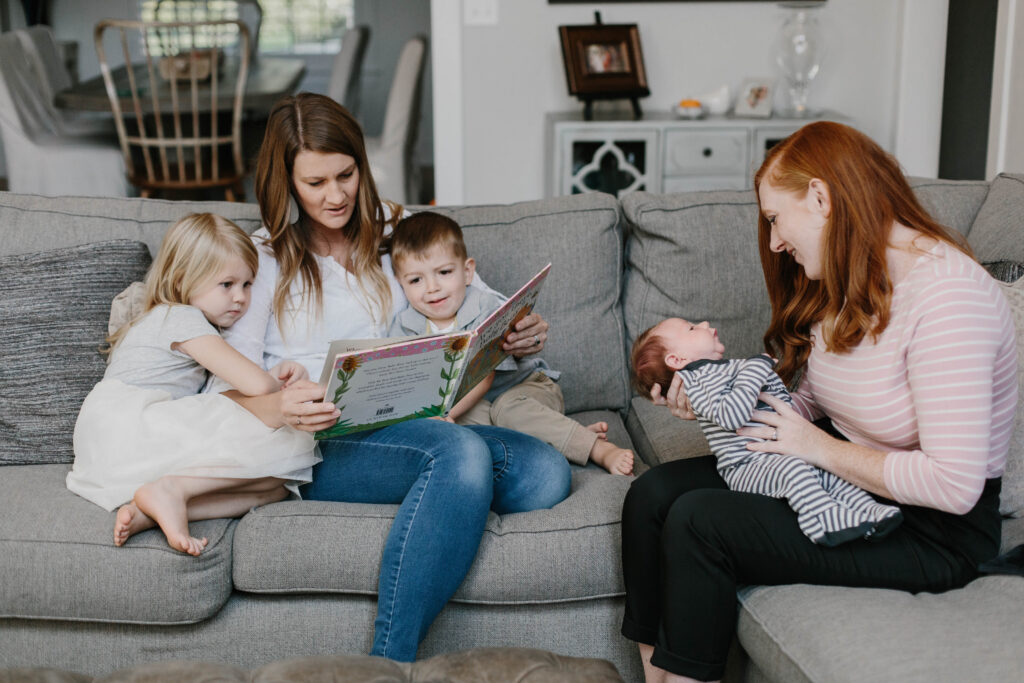 Adult reading a picture book to two young children while another adult holds a newborn on a living room couch